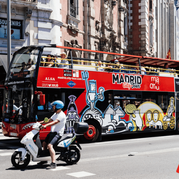 Red, wrapped double decker bus filled with people driving on the road with a man on a white moped by them.