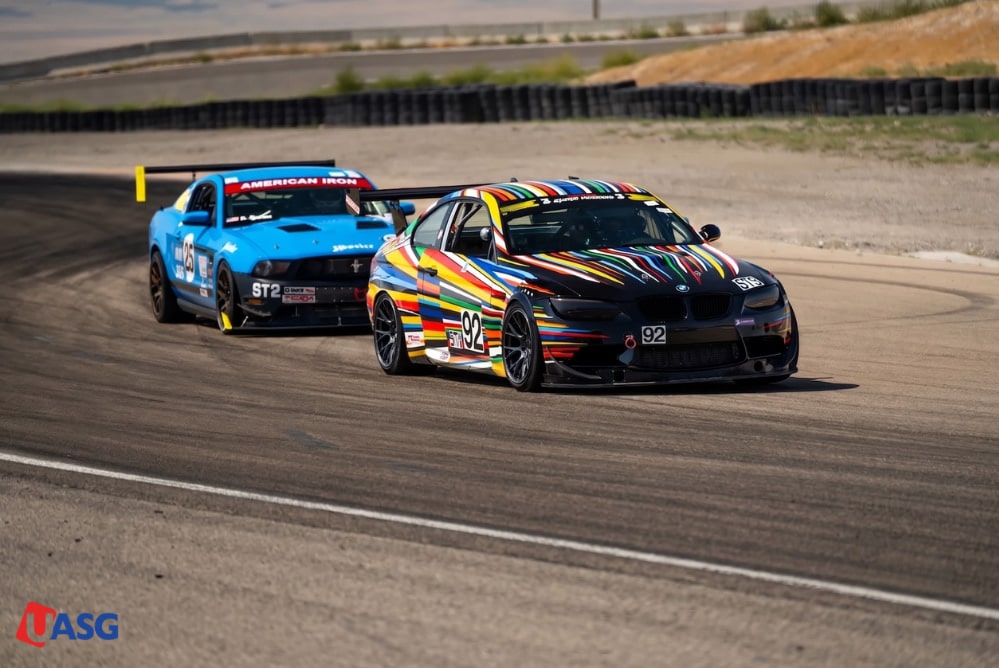 Two vinyl-wrapped race cars, a colorful BMW leading a blue Mustang, on a racetrack.